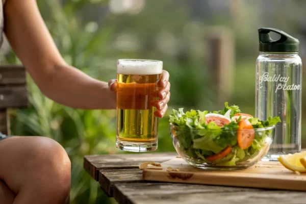 A lifestyle image showing a person enjoying a glass of golden beer in a relaxed, natural setting—perhaps on a patio or at a picnic table—alongside a fresh salad and a water bottle, suggesting balance and moderation.